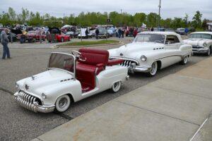 A classic white car towing a miniature matching car with red seats at a car show.