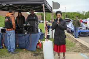 A woman speaks into a microphone outdoors near a tent and sound equipment.