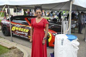 A woman in a red dress poses in front of a race car at an outdoor event.