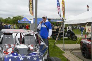 Man in blue shirt standing next to a race car at an outdoor event.
