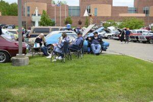 People relaxing on lawn chairs near classic cars at an outdoor event.
