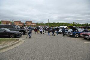 People gather at an outdoor car event with cars lined up on both sides.