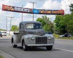 Vintage gray pickup truck driving under a welcome banner on a sunny day.