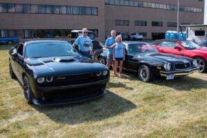 Two men standing between a Dodge Challenger and a classic car at an outdoor car show.