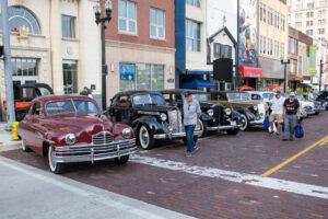 A man stands near a row of vintage cars parked on a city street.