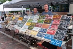 Two men proudly display a colorful collection of vintage license plates at an outdoor market.