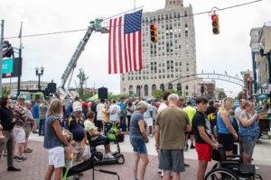 Crowd gathered under a large American flag in a city square with historic buildings.