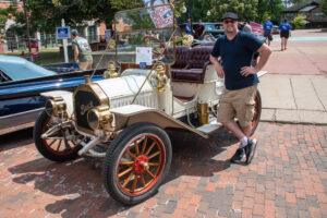 A man stands next to a vintage cream-colored car with red wheels on a brick road.