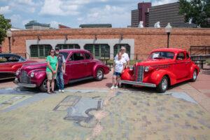 Two people stand beside classic vintage cars in an urban outdoor setting.
