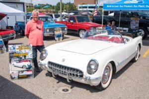 A man stands proudly beside his classic white convertible car at an outdoor event.