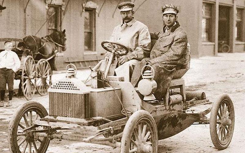 Two men driving an early vintage car.
