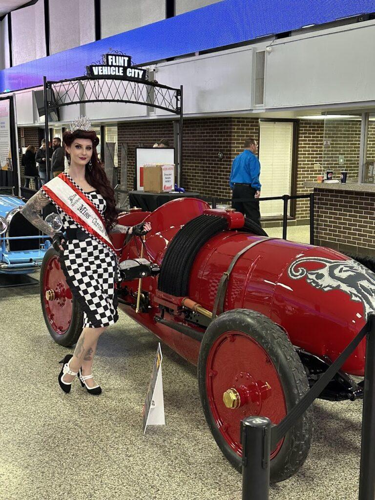 Crowned woman in checkered dress beside red racer